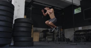 Spor salonunda çapraz antrenman yapan üstsüz, beyaz bir adamın ön görüntüsü, hafif engellerin üzerinden atlamak, ağır çekim...