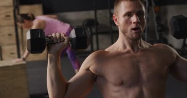 Spor salonunda oturan ve halterleri kafasının üstüne kaldıran, arka planda egzersiz yapan, ağır çekimde spor yapan üstsüz, beyaz bir adamın ön görüntüsü.