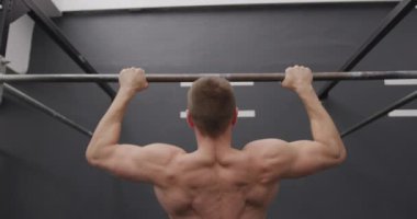 Spor salonunda antrenman yapan, barın üstünde barfiks çeken, ağır çekimde barfiks çeken üstsüz beyaz bir adamın arka görüntüsü.