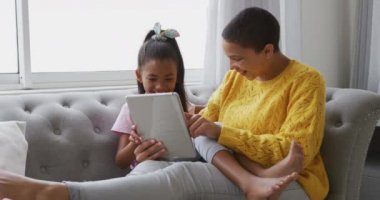 Gülümseyen bir Afro-Amerikalı kadın ve küçük kızı oturma odasında, tablet bilgisayar kullanarak rahat bir şekilde yaslanıyorlar ve gülümsüyorlar, yavaş çekim.
