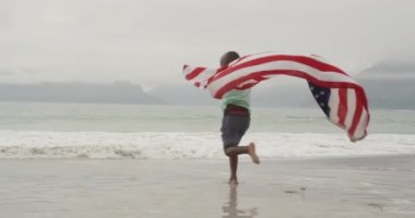 Sırtında Amerikan bayrağıyla sahile doğru koşan Afrikalı genç bir çocuğun arka görüntüsü, ağır çekimde.