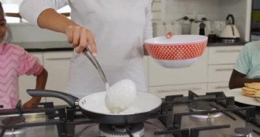 Afrika kökenli Amerikalı bir kadının mutfağa girip küçük kızı ve oğlu tarafından izlenen bir kepçeyle krep karışımını kızartma tavasına koymasının ön tarafı.