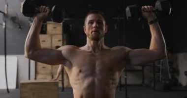 Spor salonunda antreman yapan üstsüz, beyaz bir adamın ön görüntüsü, arka planda egzersiz yapan bir kadın ve kafasının üstünde dambıllar var, ağır çekimde.