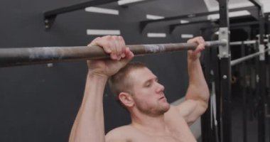 Spor salonunda antrenman yapan üstsüz, beyaz bir adamın barın üstünde barfiks çekişini.