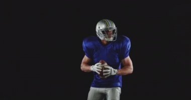 Kafkas asıllı Amerikan futbolcusunun takım üniforması, pedleri ve kaskı var elinde futbol topu, etrafa bakıyor ve blöf yapıyor, ağır çekim.