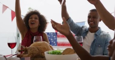 Afrika kökenli Amerikalı çok nesilli bir ailenin Bağımsızlık Günü yemeği için bayraklarla süslenmiş bir yemek masasının etrafında oturup bayrakları sallayarak ve balonlarla oynayarak eğlenmesini yakından izleyeceğiz.