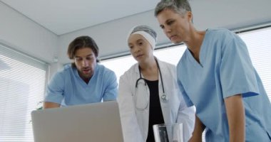 Kafkasyalı bir erkek ve kadın sağlık görevlisinin önlük giydiği ve hastanede laboratuvar önlüğü giyen, dizüstü bilgisayarın etrafında duran ve ona ağır çekimde bakan melez bir kadın sağlık görevlisinin ön görüntüsü.