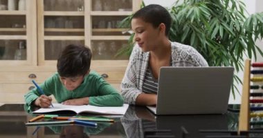 Afrika kökenli Amerikalı bir kadın ve melez oğlu, mutfakta oturmuş dizüstü bilgisayar kullanıyor ve oğlunun ödevini ağır çekimde yapmasını seyrediyor.