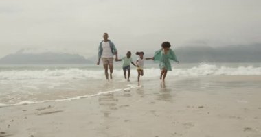 Genç oğulları ve kızlarıyla el ele tutuşan, deniz kenarında koşan Afrikalı Amerikalı bir çiftin ön görüntüsü, yavaş çekim.