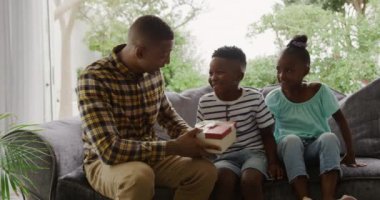 Afro-Amerikalı bir adam ve küçük oğlu ve kızı oturma odasında bir koltukta otururken, baba oğluna hediye olarak bir hediye kutusu veriyor, ağır çekimde.
