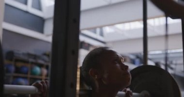 Spor kıyafetleri giymiş, spor kıyafetlerini giymiş spor salonunda eğitilmiş omuzlarında halterle ağırlık kaldıran ve kürsüde onları yavaşça değiştiren bir kadının yan görüntüsü.