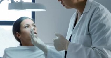 Kafkasyalı bir kadının lambanın altında otururken türban ve cerrahi eldiven takan melez bir kadın doktorun klinikte estetik yüz bakımı yaptıktan sonra yüzünü incelemesi ön planda.