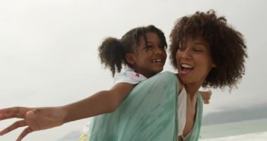 Yan görüntüde, genç kızını sırtında taşıyan ve gülümseyen, ağır çekim gülümseyen Afro-Amerikalı bir kadın var.