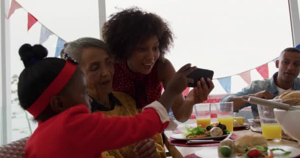 彼女の祖母と母親に家族の食事中にスマートフォンで写真を見せながら家で夕食のテーブルに座っている若いアフリカ系アメリカ人の女の子のサイドビュー — ストック動画