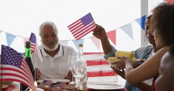 私たちと一緒に飾られたディナーテーブルの周りに家に座っているアフリカ系アメリカ人の多世代の家族の側面図独立記念日のお祝いの食事 食べ物のボウルを保持し 笑顔と旗を振って スローモーション — ストック動画