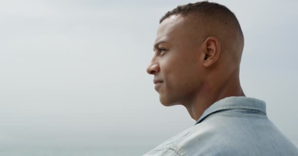 一个留着短发 穿着T恤和斜纹棉布衬衫的非裔美国人站在海滩上 转向镜头 慢吞吞地微笑着 侧视近景 — 图库视频影像