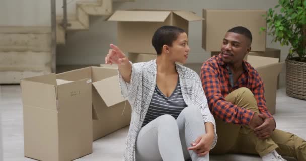 ボックスに囲まれた新しい家の床の上に座っているアフリカ系アメリカ人のカップルのフロントビューでは 物事を置くためにどこに スローモーションを議論 — ストック動画
