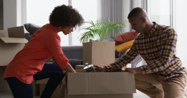 一对非洲裔美国人夫妇跪在客厅的地板上 笑着从纸板箱中打开一只咖啡壶 四周都是包装箱 动作缓慢 — 图库视频影像