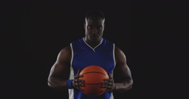 Porträt eines muskulösen afrikanisch-amerikanischen Basketballspielers in den Mannschaftsfarben, der einen Basketball in zwei Händen vor sich hält, ihn ansieht und in die Kamera blickt, Zeitlupe