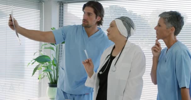 在一家医院的办公室里 一个身穿洗涤剂的白人男女医护人员和一个身穿实验室外套的混血女性医护人员站在一起 讨论着一个男人拿着的X光照片的镜头在众目睽睽之下 — 图库视频影像