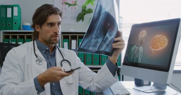 一名身穿实验室外套的白人男医生坐在一家医院的办公室里 手持X光片 用智能手机打电话 — 图库视频影像