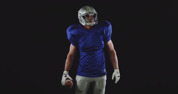Pohled zepředu na bělocha amerického fotbalisty v uniformě, polštářcích a helmě, jak stojí a drží míč, zpomalený pohyb