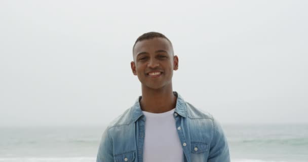 Tシャツとデニムのシャツを着たアフリカ系アメリカ人男性の肖像画で 彼の後ろの海とビーチに立って カメラに笑顔 スローモーション — ストック動画