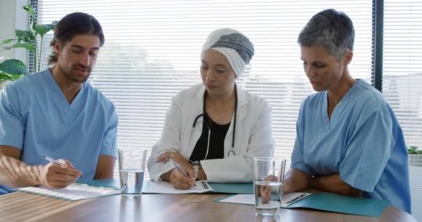 研究室のコートを着た白人男性と女性の医療従事者の正面図病院のオフィスのテーブルに座って話をしたりメモをしたり — ストック動画