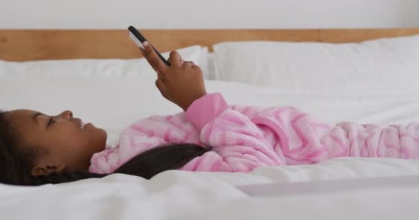ベッドの上に寝そべってスマートフォンを使って笑顔でゆっくりとした動きをしているアフリカ系アメリカ人の少女の姿が見えます — ストック動画