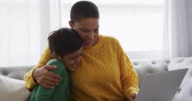 Gülümseyen bir Afro-Amerikalı kadın ve melez oğlunun oturma odasında bir koltukta oturup dizüstü bilgisayarını birlikte kullanmaları ve kucaklaşmaları.