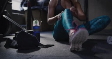 Spor kıyafetleri giyen, spor salonunda çapraz antrenman yapan, yerde oturup, esneyen, akıllı telefon kullanan, havlusunu ve su şişesini alıp uzaklaşan atletik bir kadının ön görüntüsü.