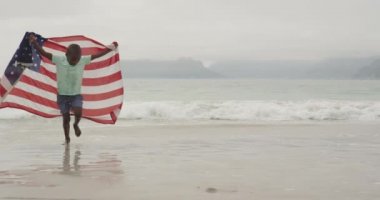 Sahilde koşan genç Afrikalı Amerikalı bir çocuğun ön görüntüsü. Omzuna Amerikan bayrağı asılmış, ağır çekimde.