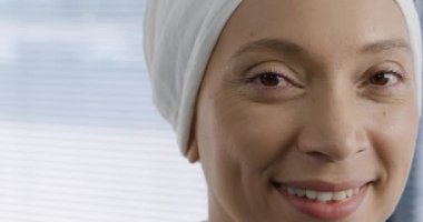 Hastanedeki bir ofisin penceresinin önünde kameraya gülümseyen türban takan melez bir kadın sağlık görevlisinin kafa görüntüsü.