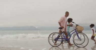 Side view of a smiling African American couple wheeling bicycles along a beach by the sea with their young daughter and son running around them, slow motion
