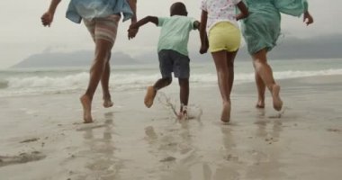 Küçük oğulları ve kızlarıyla el ele tutuşan, kumsalda koşan Afrikalı Amerikalı bir çiftin düşük açılı görüntüsü, yavaş çekim.