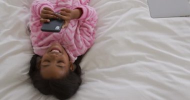 Genç Afro-Amerikan bir kızın evde olduğu yüksek açılı bir manzara. Yatakta sırt üstü uzanmış, tulum giyiyor, akıllı telefon kullanıyor ve gülümsüyor, ağır çekim.