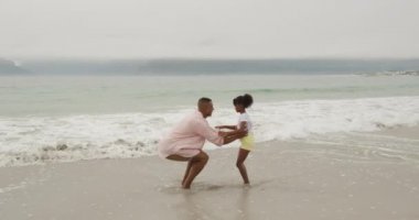 Sahilde duran Afrikalı Amerikalı bir adamın yan görüntüsü. Genç kızını havaya fırlatıyor ve onu yavaş çekimde yakalıyor.