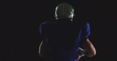 Kafkas asıllı Amerikalı bir futbolcunun takım üniforması, pedleri ve kaskı var, bir elinde futbol topu tutuyor ve iki kolunu da zaferle kaldırıyor ve kameraya doğru dönüyor. Yavaş çekim