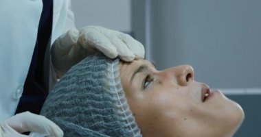 Bir doktor klinikte alnına iğne yaparken, beyaz bir kadının şapkasını yere sererken yan görüntüsü.