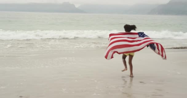 一个年轻的非洲裔美国女孩在海滩上跑向大海 肩上披着一面美国国旗 动作缓慢的后视镜 — 图库视频影像