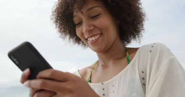 一个非洲裔美国女人用智能手机在海滩上散步 慢吞吞地笑着的侧视图 — 图库视频影像