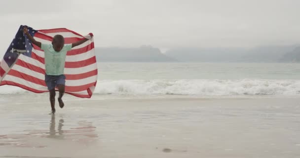 一个年轻的非洲裔美国男孩在海滩上冲向镜头 肩膀上挂着一面美国国旗 动作缓慢 — 图库视频影像