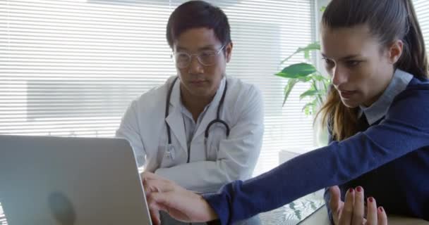 ノートパソコンの画面を指し 病院のオフィスで相談中にアジアの男性医師と話す机に座っている白人女性患者のフロントビュー — ストック動画