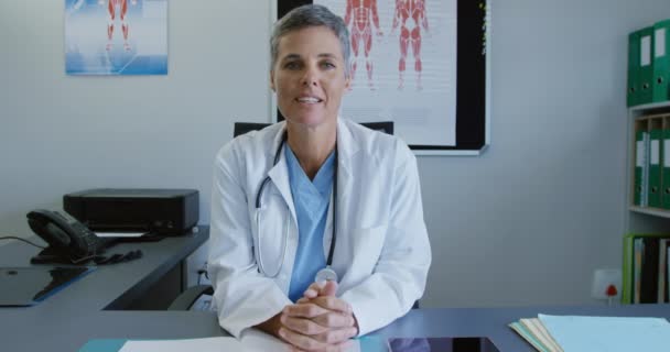 研究室のコートを着た白人女性医師の正面図カメラと話す医療室の机の後ろに座って — ストック動画