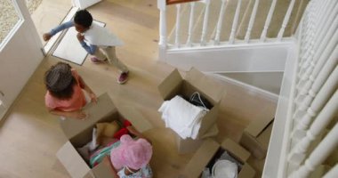 Afrika kökenli Amerikalı bir annenin yeni evinin koridorunda diz çöküp küçük oğlu ve kızıyla birlikte yardım ve oyun oynadığı bir kutu şapkayı açarken görüntüsü.