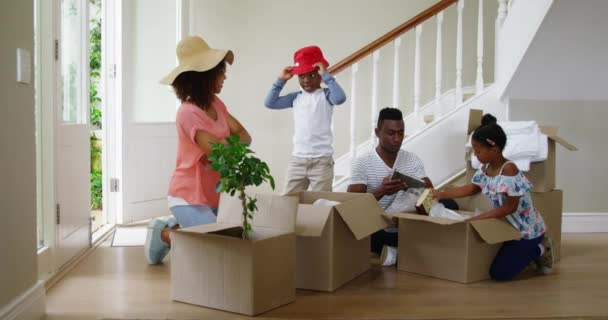 新しい家の廊下で一緒に箱を開けるアフリカ系アメリカ人の家族と彼らの若い息子と娘の正面図 帽子を試着する母親と息子 スローモーション — ストック動画