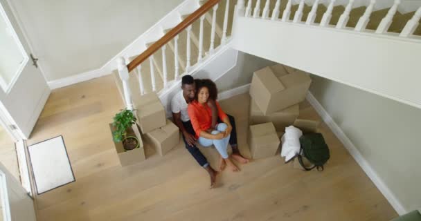 彼らの新しい家の廊下に箱の山の前に一緒に床に座っているアフリカ系アメリカ人のカップルの高い角度のビューは しばらくの間 移動中に休息し 周りを見回し スローモーション — ストック動画