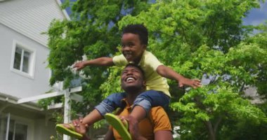 Bahçede gülümseyen Afrikalı Amerikalı bir adamın ön görüntüsü. Küçük oğlu omuzlarında oturuyor, ayak bileklerini tutuyor ve yavaşça arkasını dönüyor.