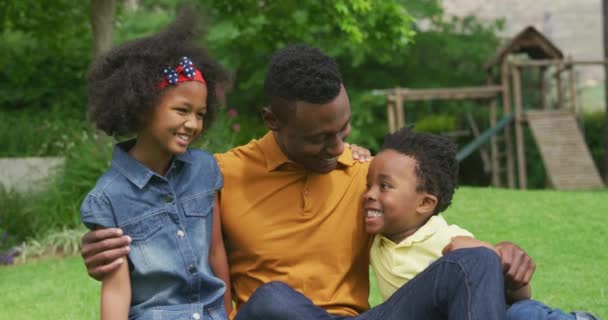 彼の幼い息子と娘の周りに彼の腕で庭に草の上に座っているアフリカ系アメリカ人の男の肖像画は すべてのカメラに笑顔 スローモーション — ストック動画