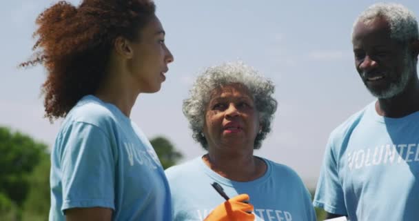 一名年轻的非洲裔美国妇女 一名男子和一对年长的夫妇站在田里 戴着手套 穿着印有志愿者字样的T恤衫 她在剪贴板上书写 并在收集垃圾前指导其他志愿者 — 图库视频影像